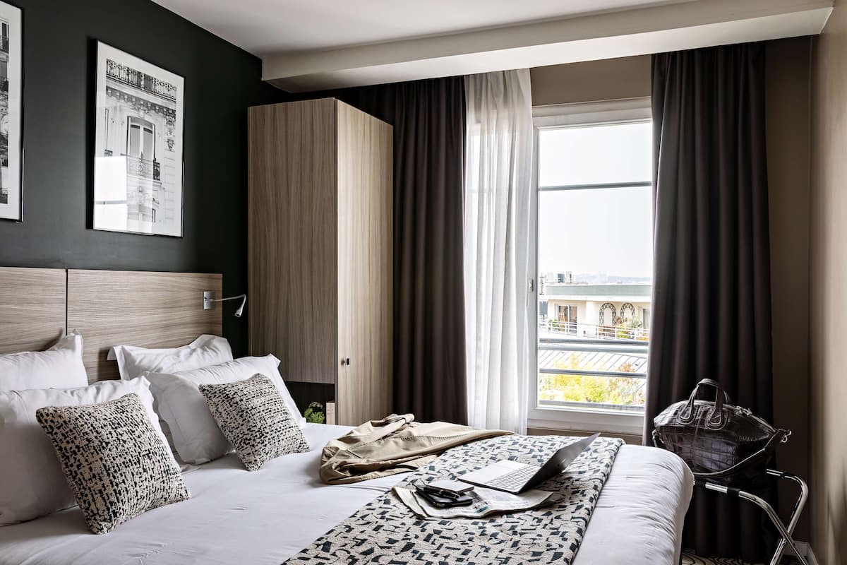Une chambre d’hôtel avec un lit, un ordinateur portable, un sac à main et une vue sur la ville grâce à de grandes fenêtres.