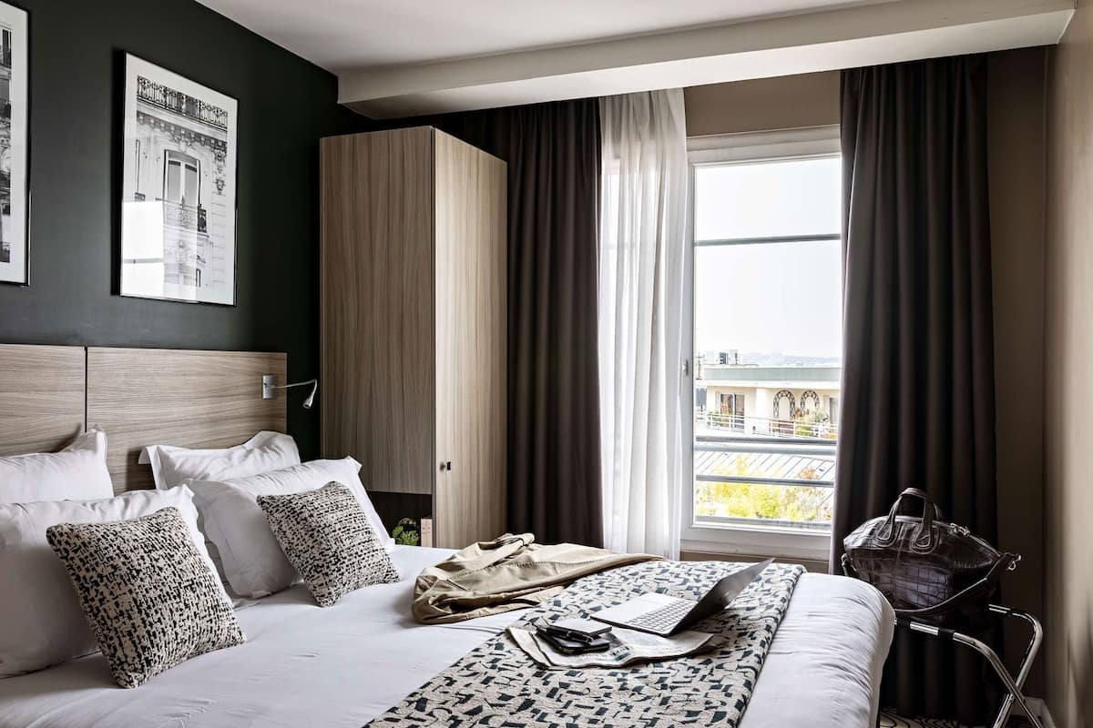 Une chambre d’hôtel comprenant un lit, un bureau, une chaise, une fenêtre avec des rideaux et une vue sur des immeubles.