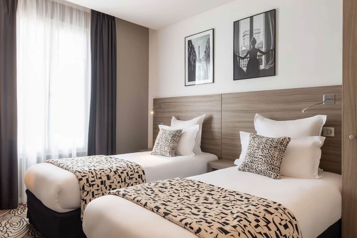 Une chambre d’hôtel avec deux lits, une grande fenêtre avec des rideaux légers et deux tableaux encadrés accrochés au mur.
