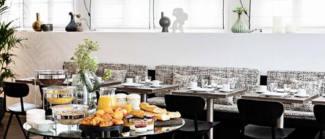 A breakfast buffet with various food items and a coffee station, set in a modern dining area with black chairs and a patterned bench.