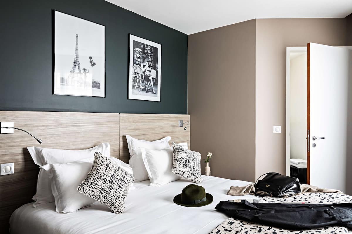Une chambre d’hôtel avec un lit, des oreillers, un chapeau, un sac à main et deux tableaux encadrés accrochés au mur.
