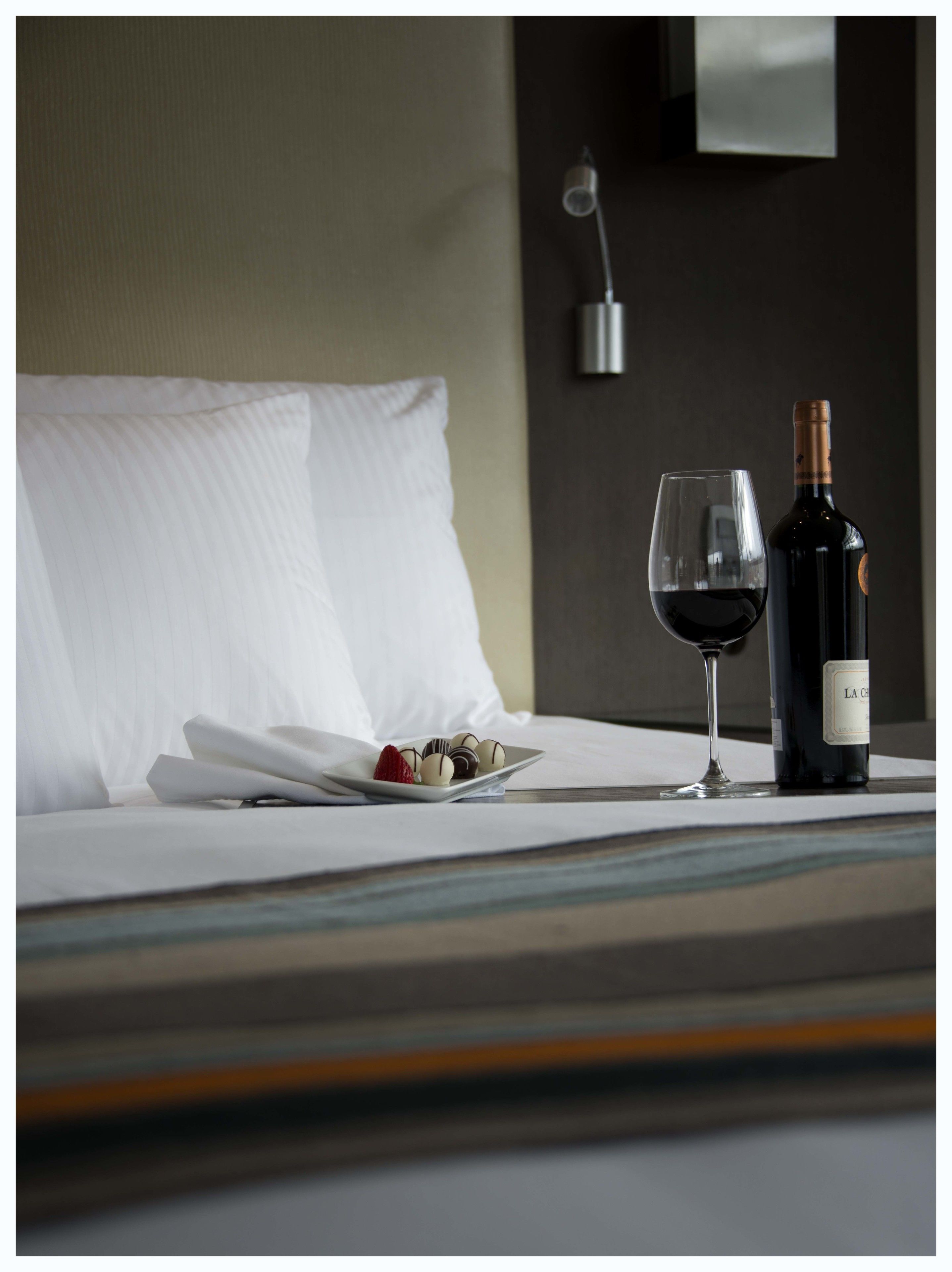 A neatly made bed with a plate of chocolates and a glass of red wine, and a bottle of wine on the bedside table.