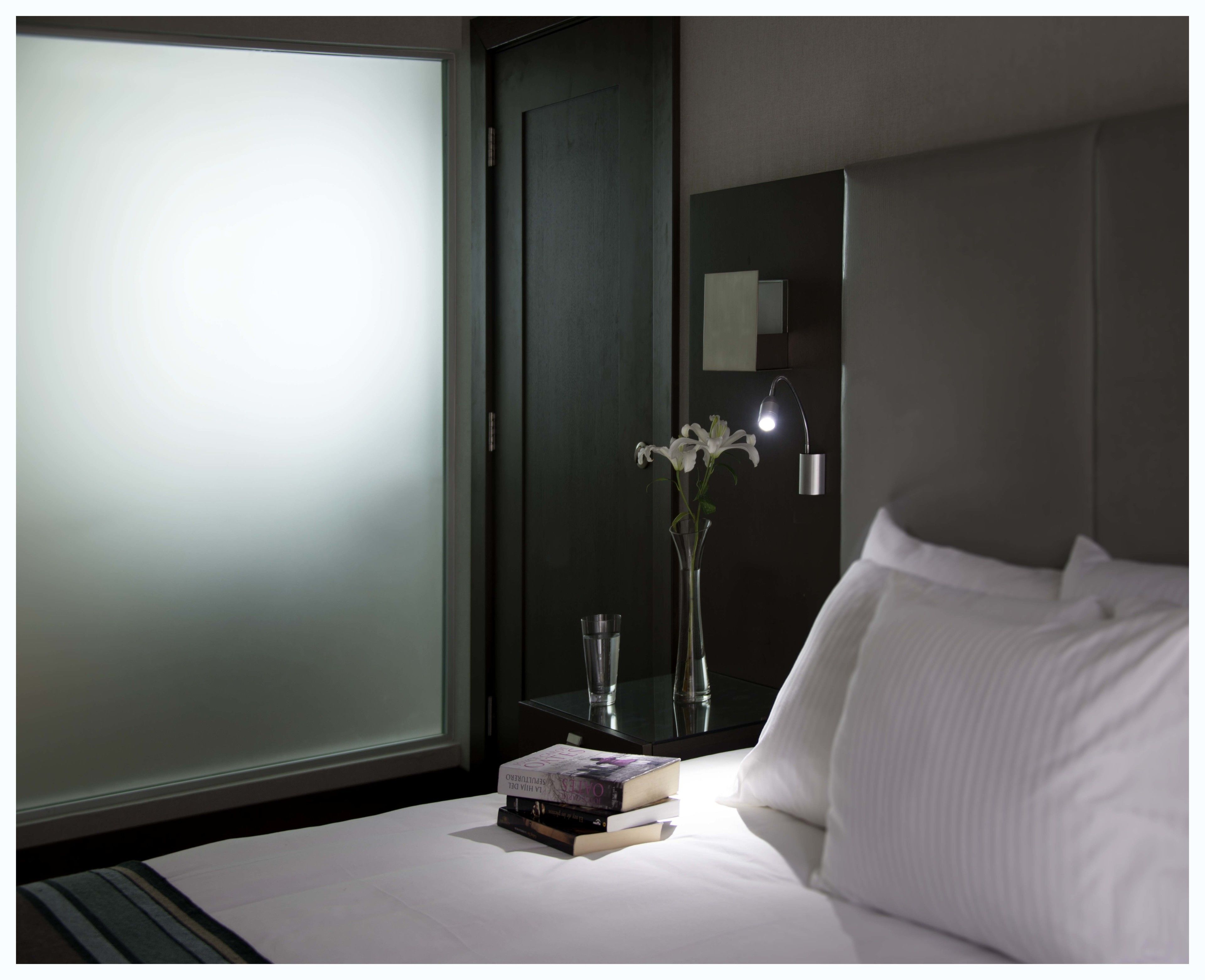 Una habitación de hotel con una cama, una mesita de noche con un libro y un vaso, un florero con flores y un espejo en la pared.