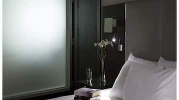 Una habitación de hotel con una cama, una mesita de noche con un libro y un vaso, un florero con flores y un espejo en la pared.