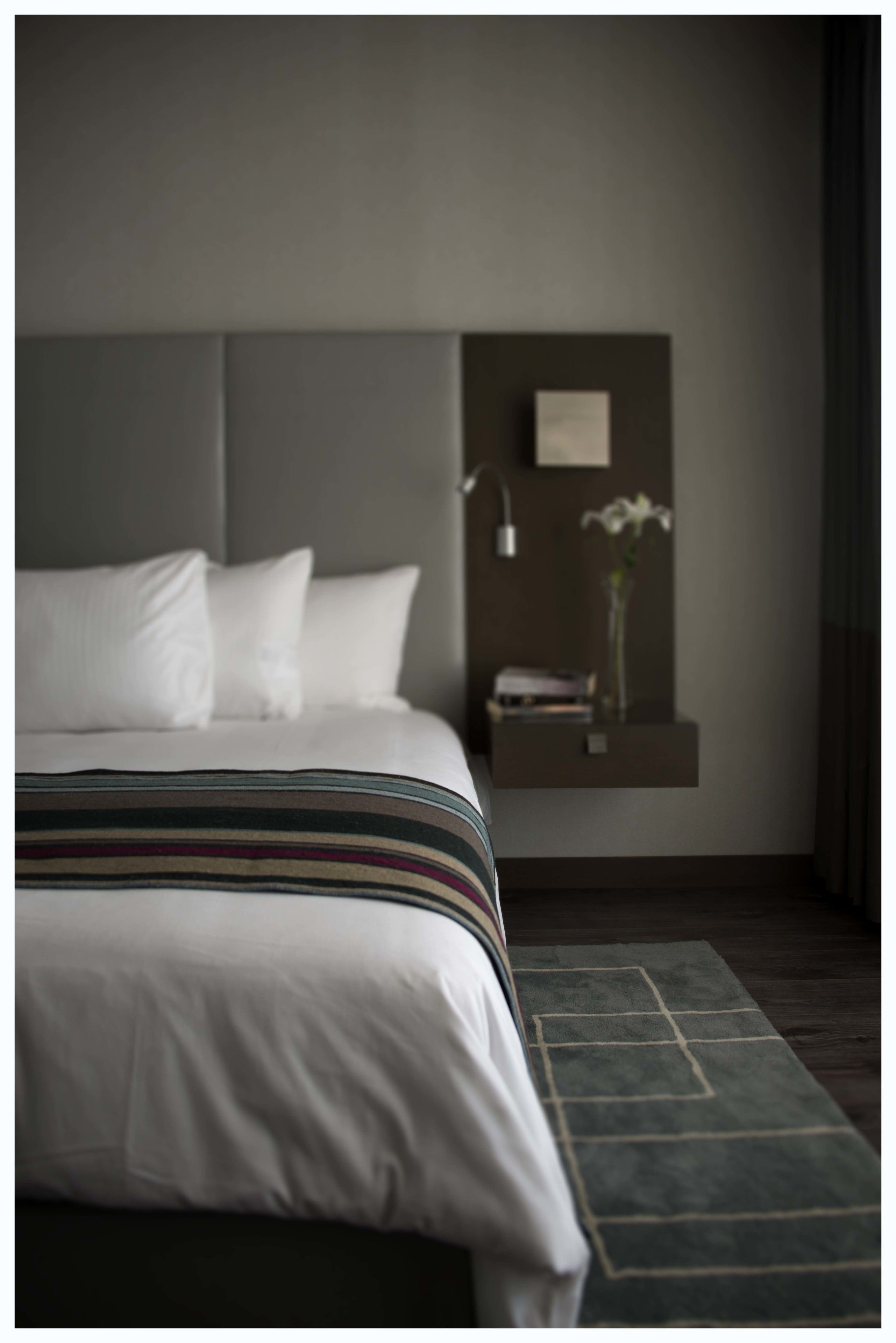 Quarto de hotel moderno com uma cama grande, uma mesa de cabeceira, um vaso com uma flor e uma prateleira fixada na parede.