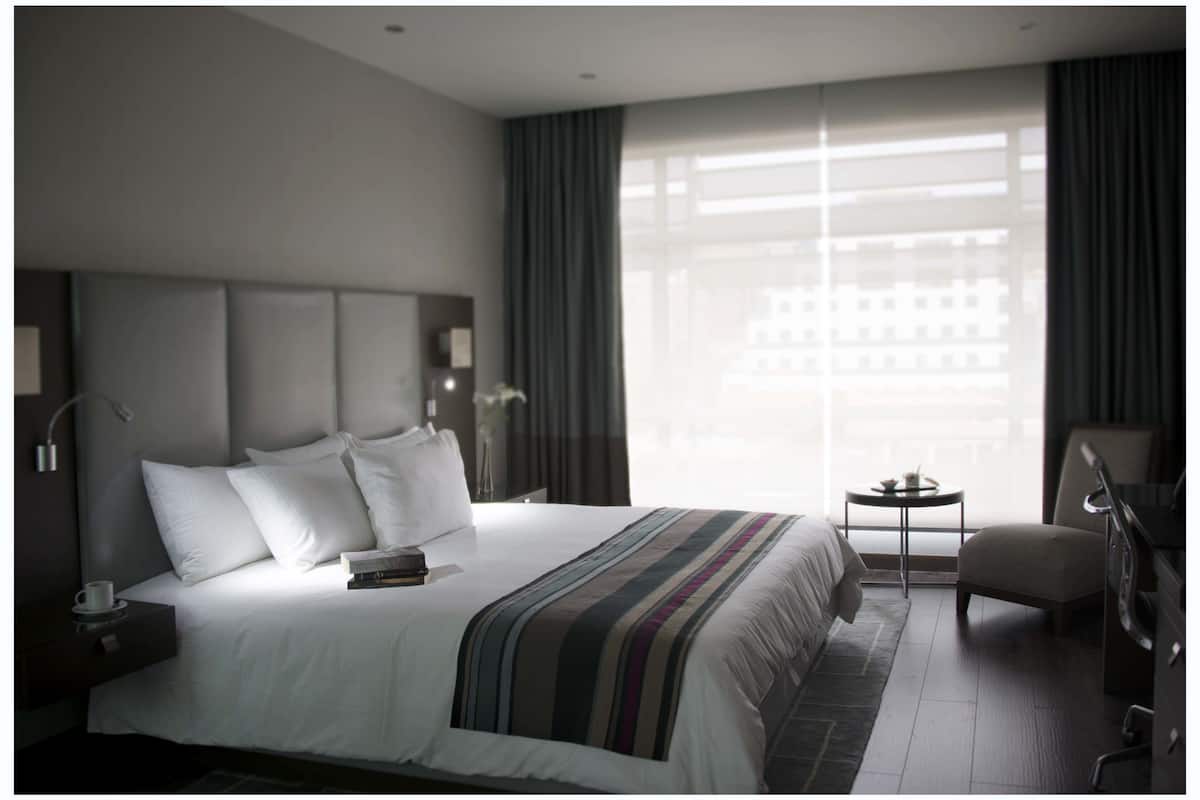 A modern hotel room with a large bed, a bedside table with a lamp, a small table with a cup, and a view of the city through large windows.