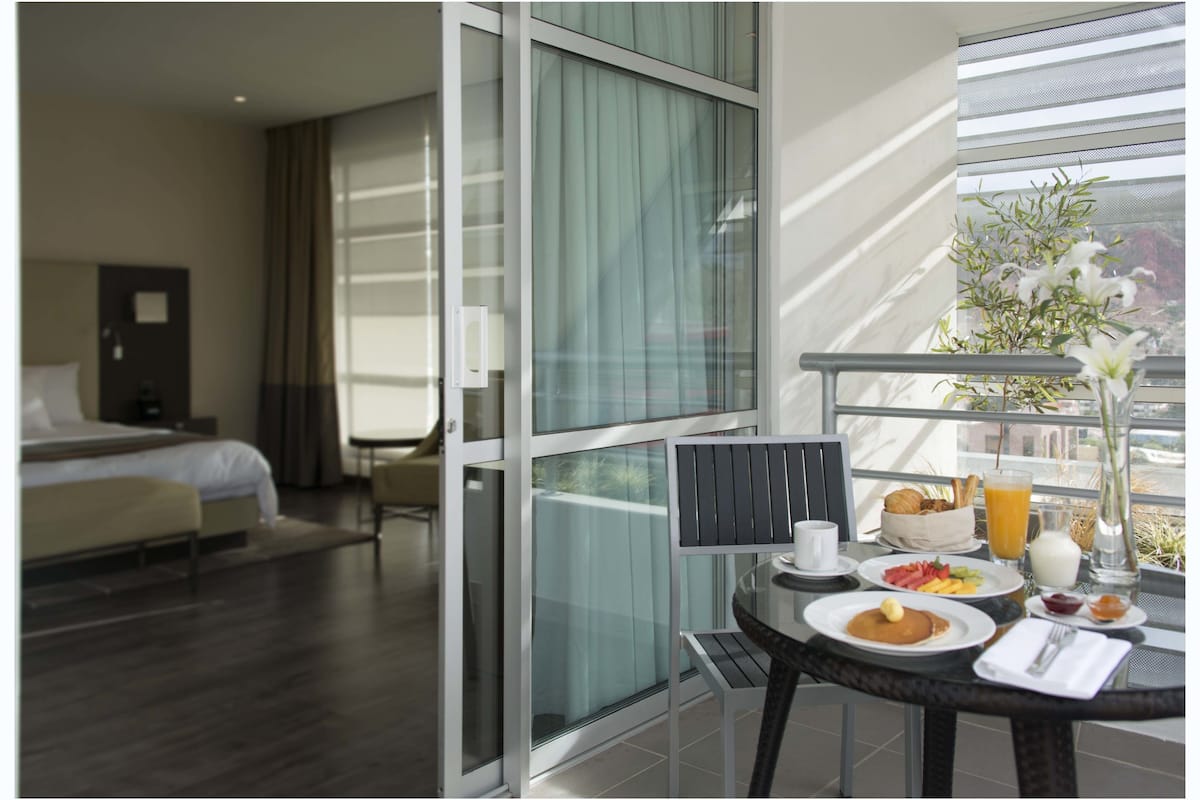 A hotel room with a bed, a chair, a balcony with breakfast items, and a view of the outdoors.