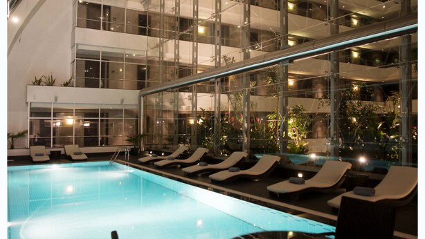 An indoor swimming pool with lounge chairs and tables, surrounded by a multi-level glass atrium with natural light and plants. - Casa Grande Hotel (La Paz)