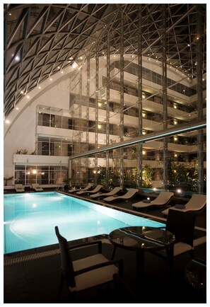 Una piscina interior con sillas de descanso y mesas, rodeada por un atrio de vidrio de varios niveles con luz natural y plantas.