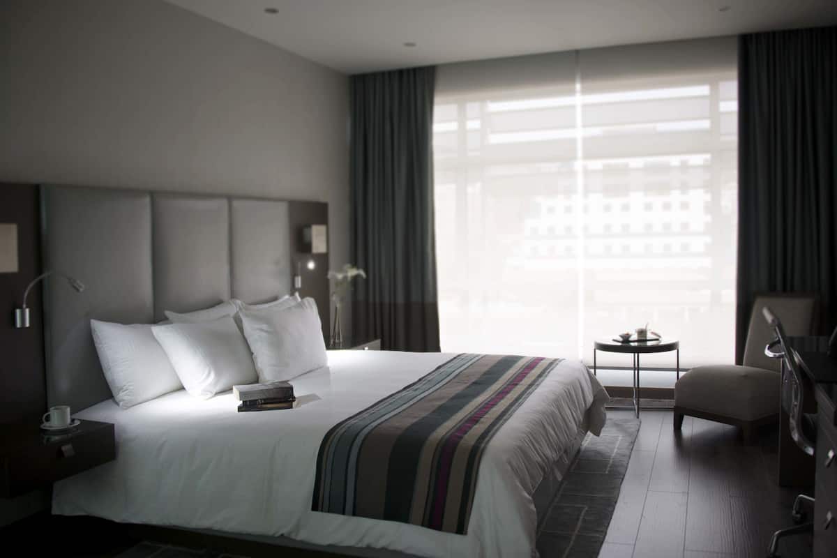 A modern hotel room with a large bed, a small table, a chair, and a view of a building through the window.