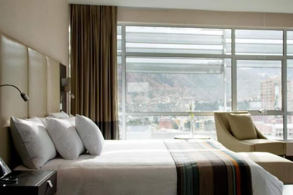 A hotel room with a large bed, a chair, a desk with a phone, and a large window offering a city view.