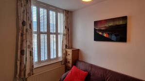 Una habitación con ventana, un radiador, un armario de madera, un sofá marrón con un cojín rojo y un cuadro en la pared.