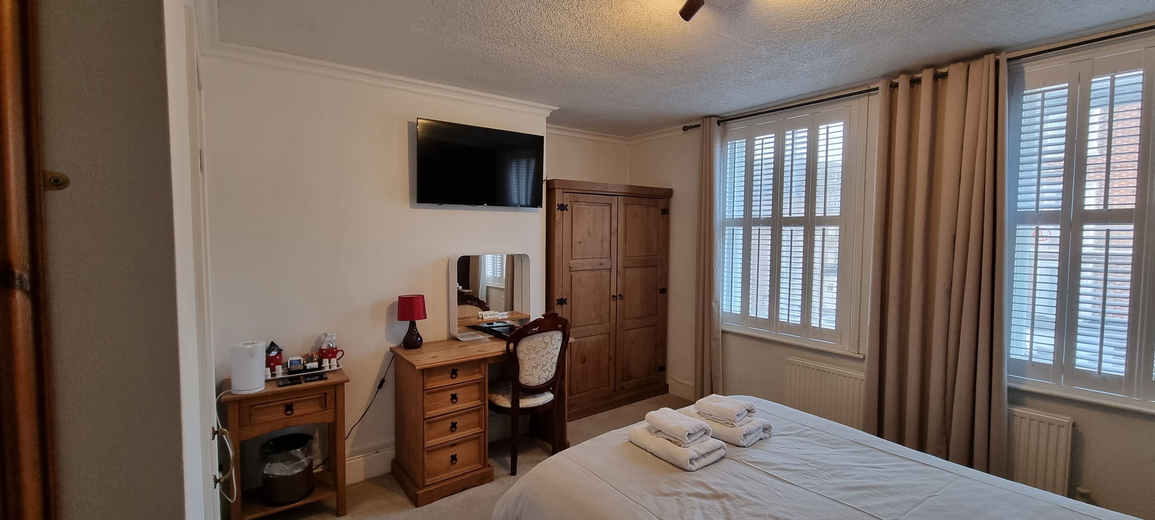 A bedroom with a bed, a desk with a lamp, a chair, a wardrobe, a television, and a window with curtains.