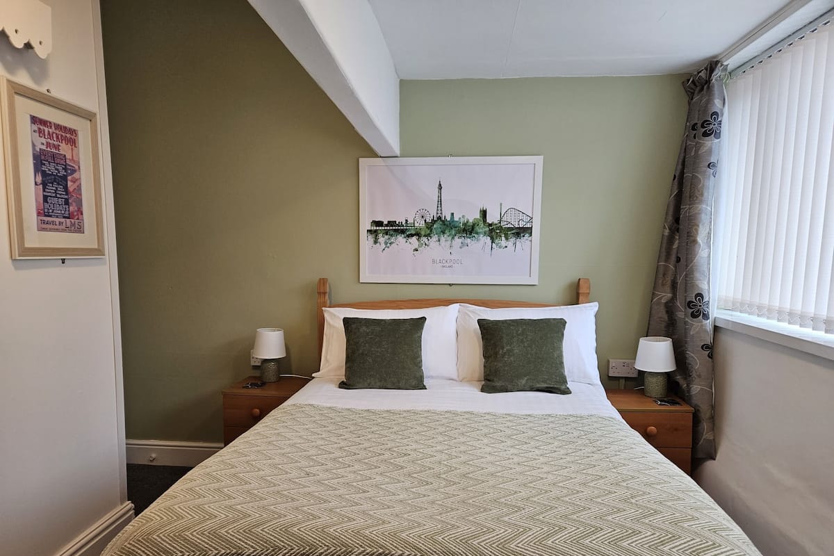 Una cama bien tendida con una colcha estampada, dos almohadas, un cuadro enmarcado con una vista urbana, una mesita de noche con una lámpara y una ventana con cortinas.