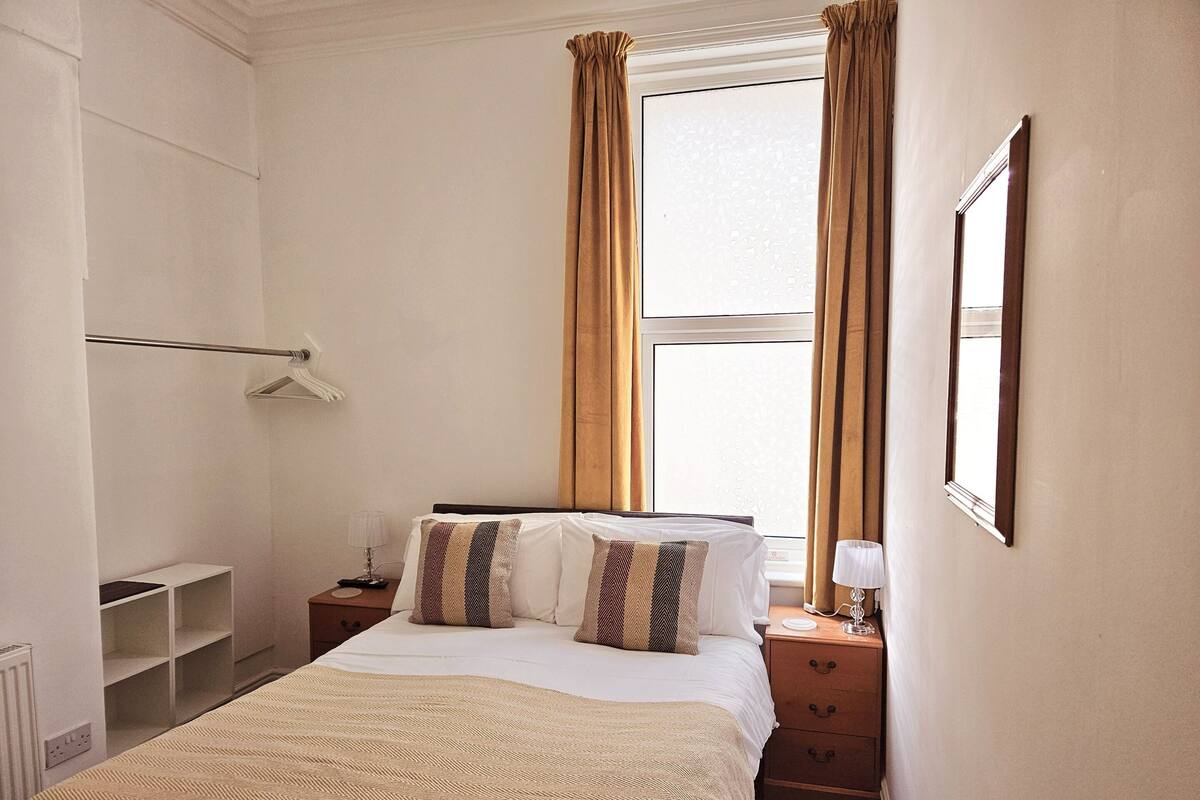 Habitación de hotel con cama, mesitas de noche, ventana con cortinas, radiador y perchero.