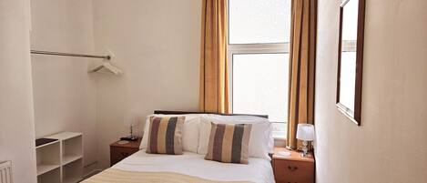 A hotel room with a bed, bedside tables, a window with curtains, a radiator, and a coat rack.