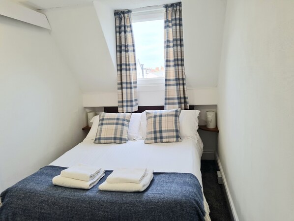 A neatly made bed with plaid pillows and folded towels, a bedside table with a lamp, and a window with plaid curtains.
