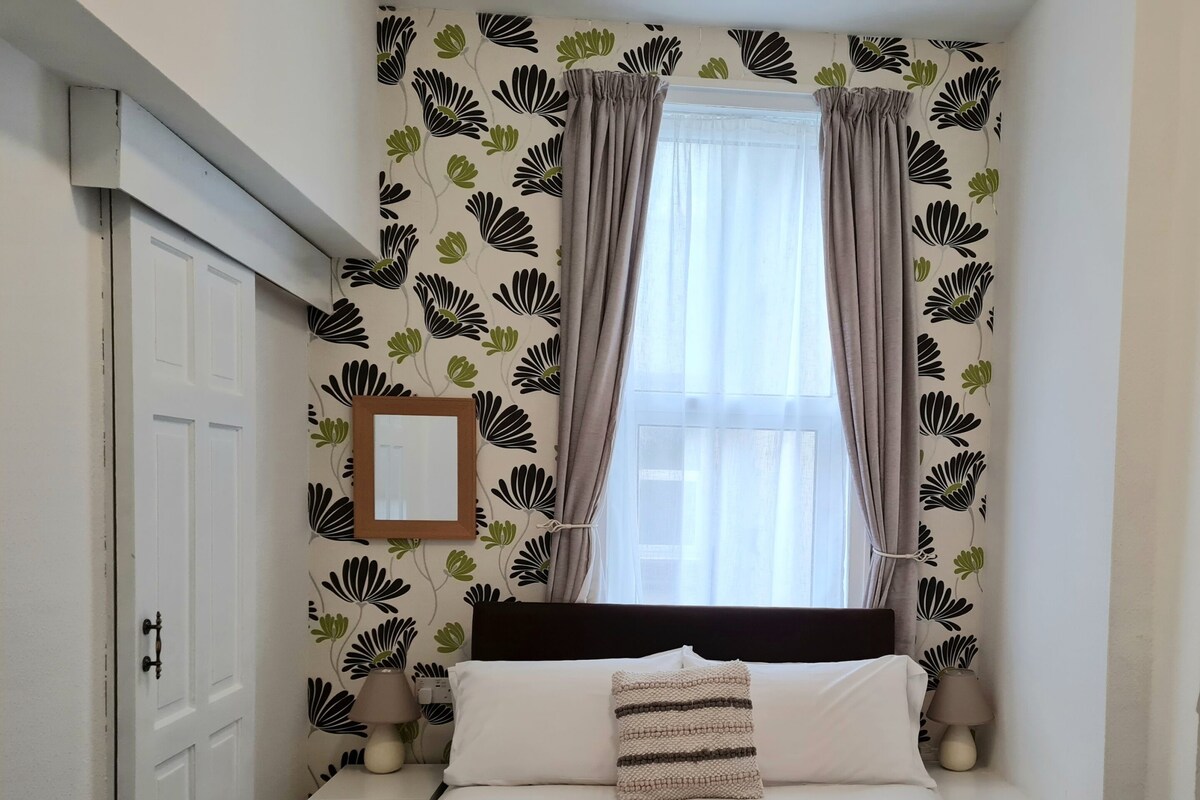 Une chambre à coucher comprenant un lit, une table de chevet, une lampe, une fenêtre avec des rideaux et un mur orné d’un motif floral.