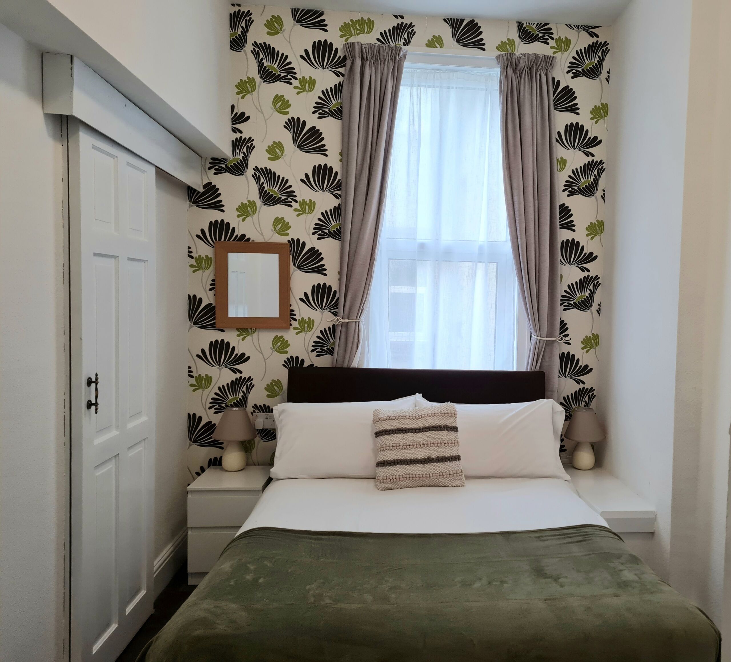 A bedroom with a bed, a nightstand, a lamp, a window with curtains, and a wall with a floral pattern.