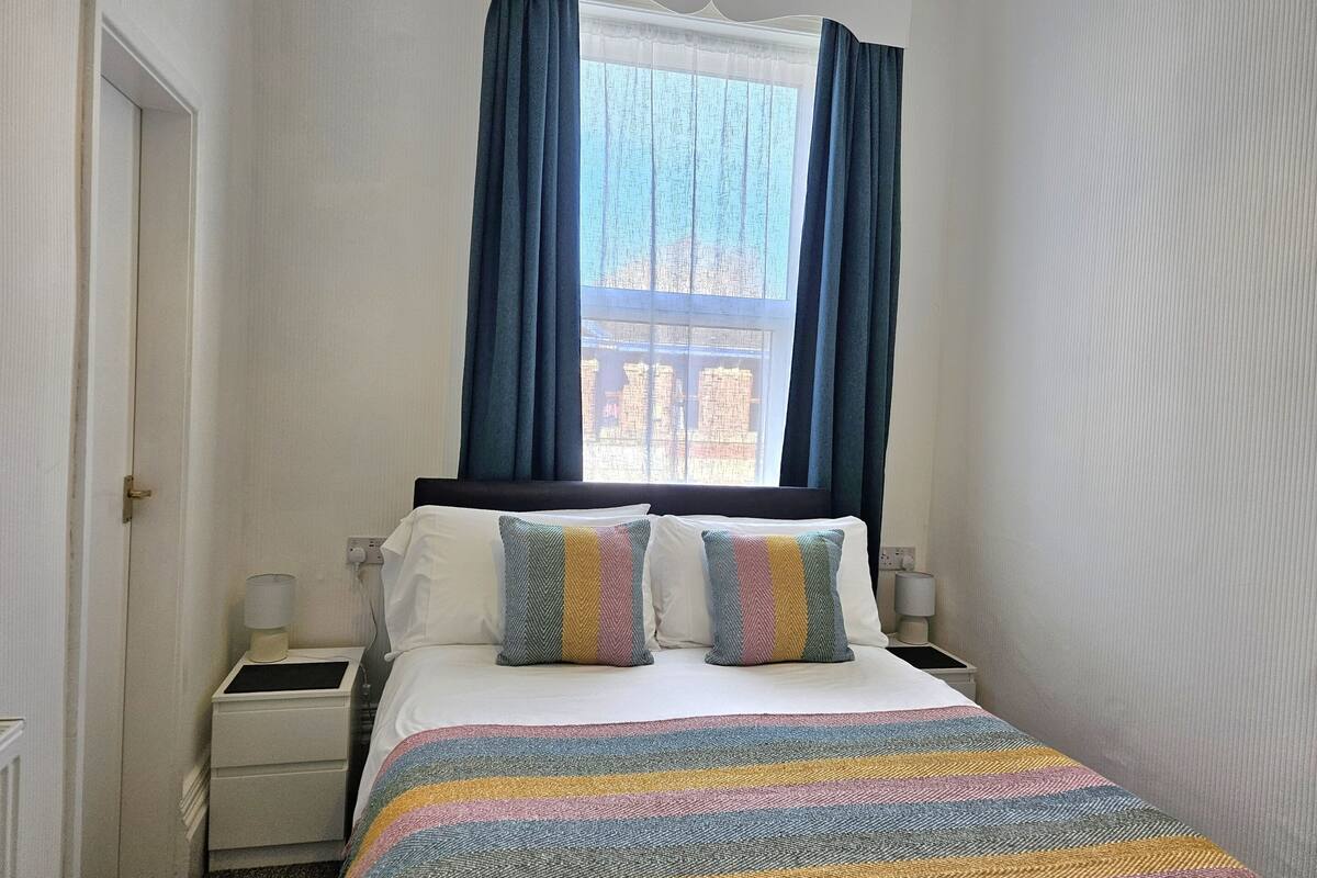 Un dormitorio con cama, dos almohadas, una mesita de noche, una lámpara y una ventana con cortinas.