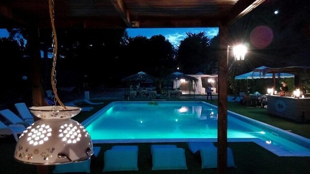 A swimming pool with a tiled deck, surrounded by lounge chairs and a covered area with tables and chairs. - Il Corbezzolo (Cupra Marittima)