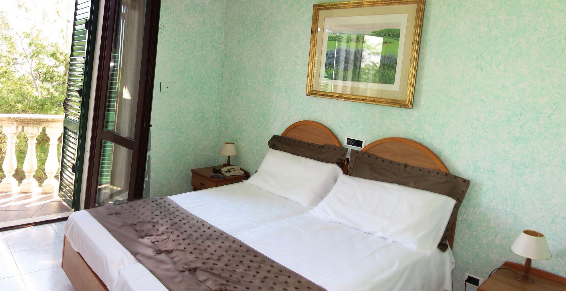 A hotel room with a bed, a nightstand, a lamp, and a window with a view of greenery.