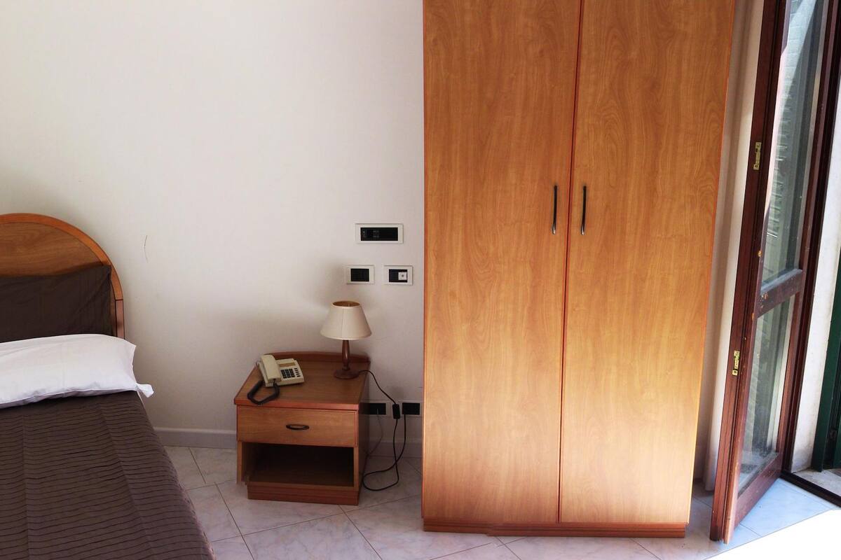 A bedroom with a bed, a nightstand with a lamp, a telephone, and a wooden wardrobe.