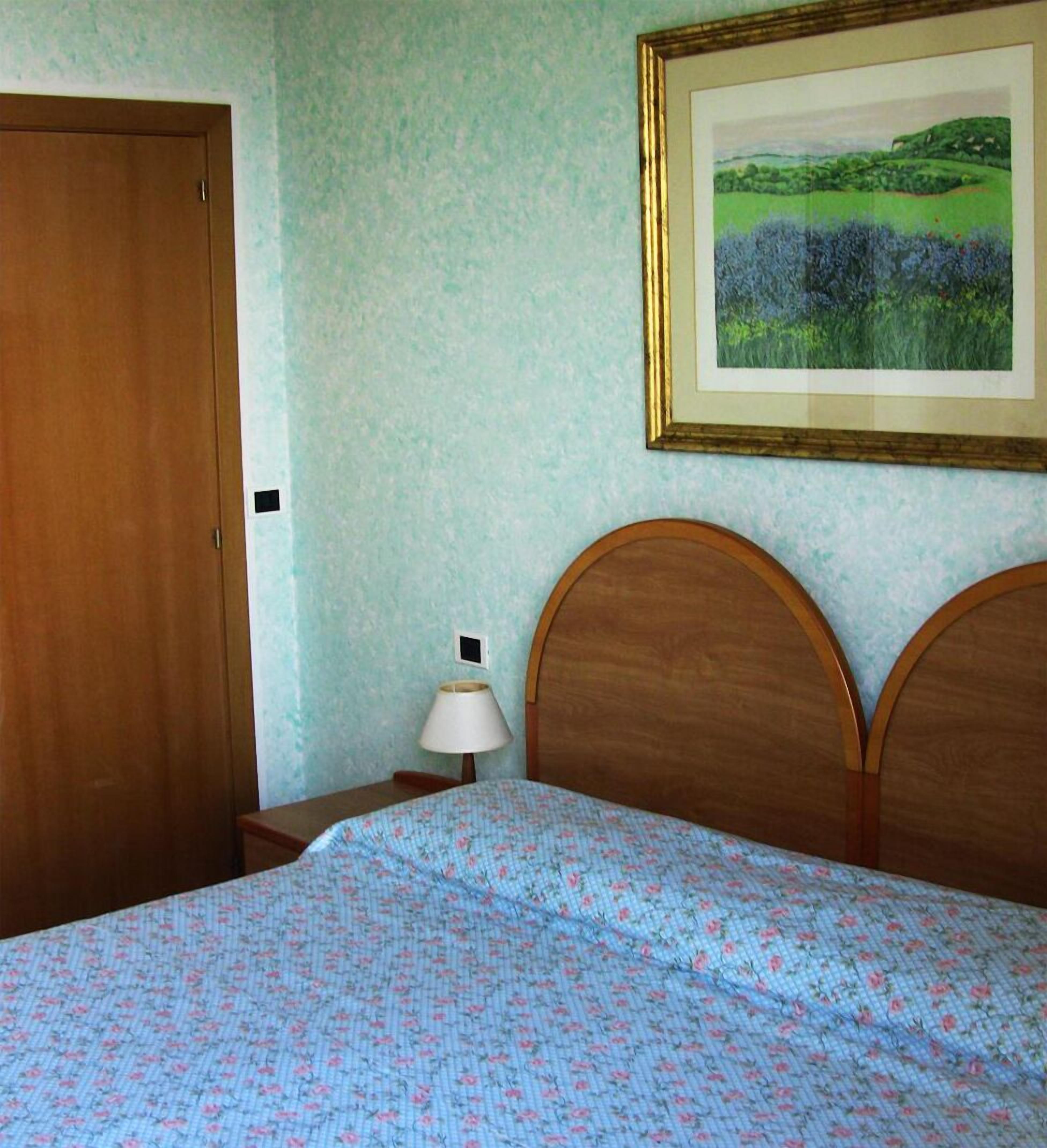 Ein Schlafzimmer mit Holztür, einem Bett mit Blumenmuster, einem Nachttisch mit Lampe und einem gerahmten Landschaftsbild an der Wand.