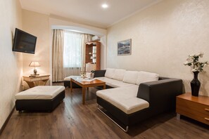 A modern hotel room with a sofa, a small table, a desk with a lamp, a TV mounted on the wall, and a window with curtains. - Odesskiy Dvorik (Odesa)