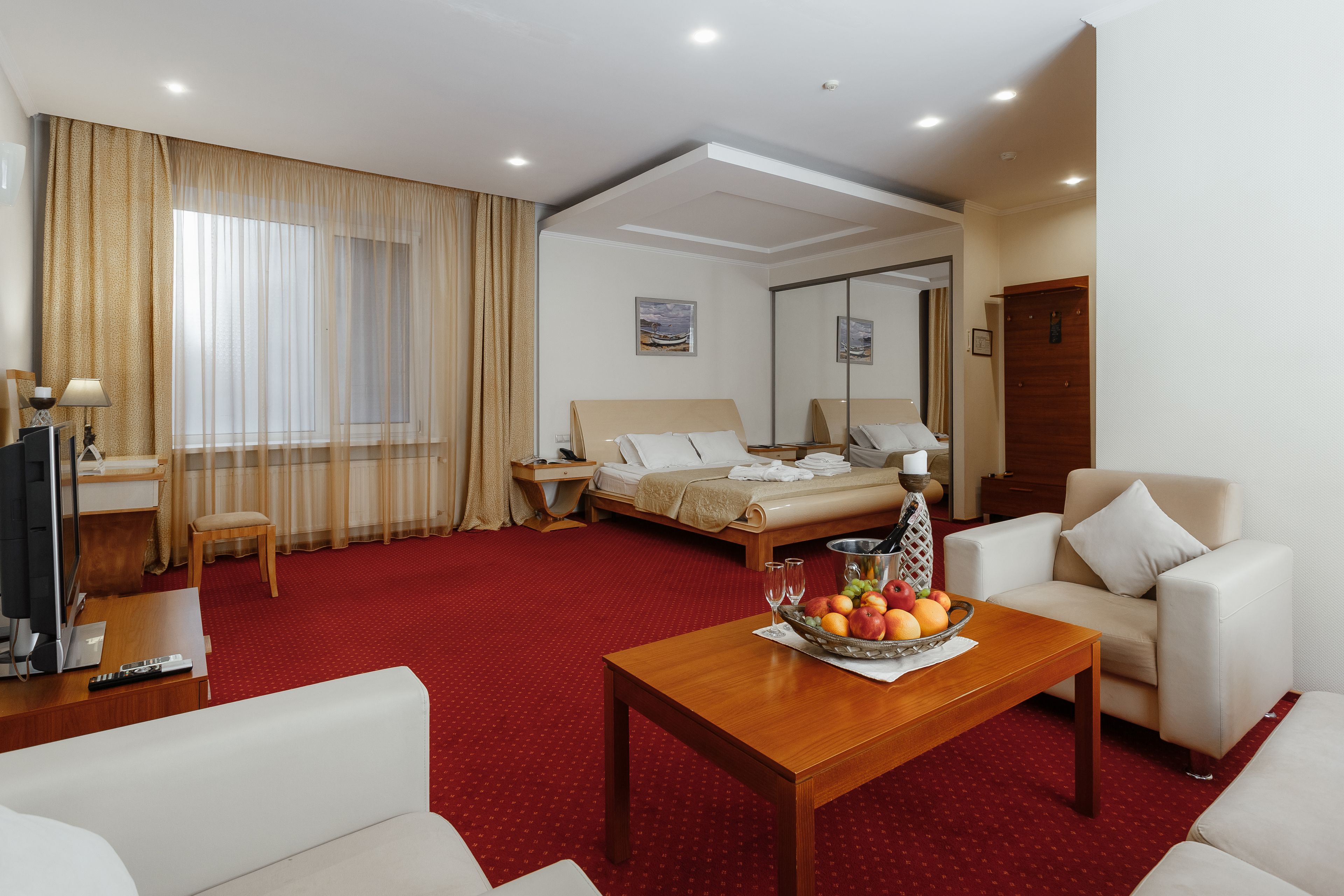 A hotel room with a bed, a sofa, a wooden coffee table with fruit, a TV, and a desk with a lamp.