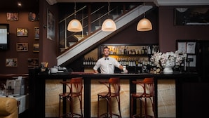 A bar area with a bartender, wooden bar stools, a staircase, and a wall of liquor bottles. - Odesskiy Dvorik (Odesa)