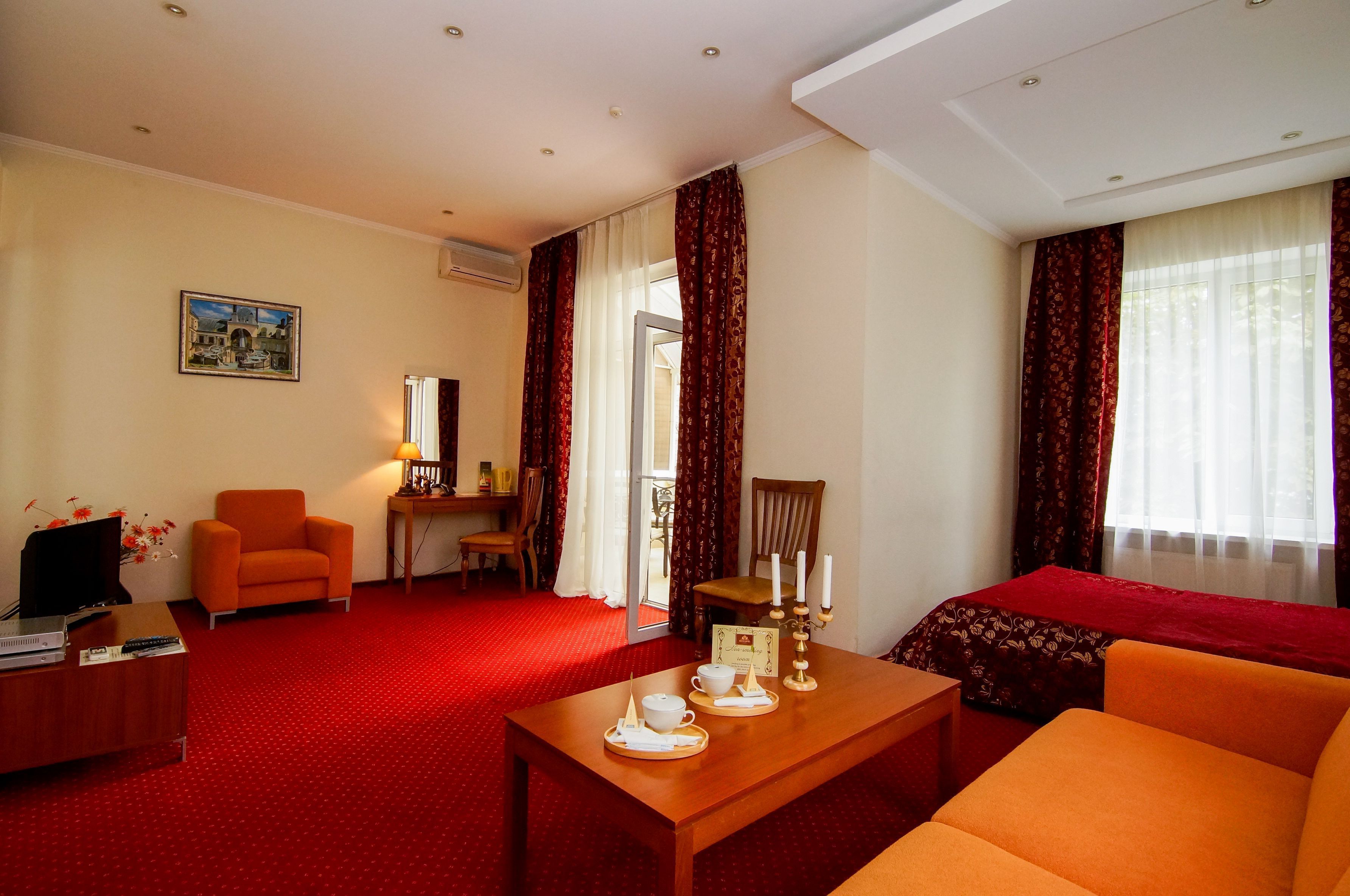 Une chambre d’hôtel avec un tapis rouge, un bureau en bois, un fauteuil orange, un canapé, une table à manger avec un service à thé et une grande fenêtre avec des rideaux.