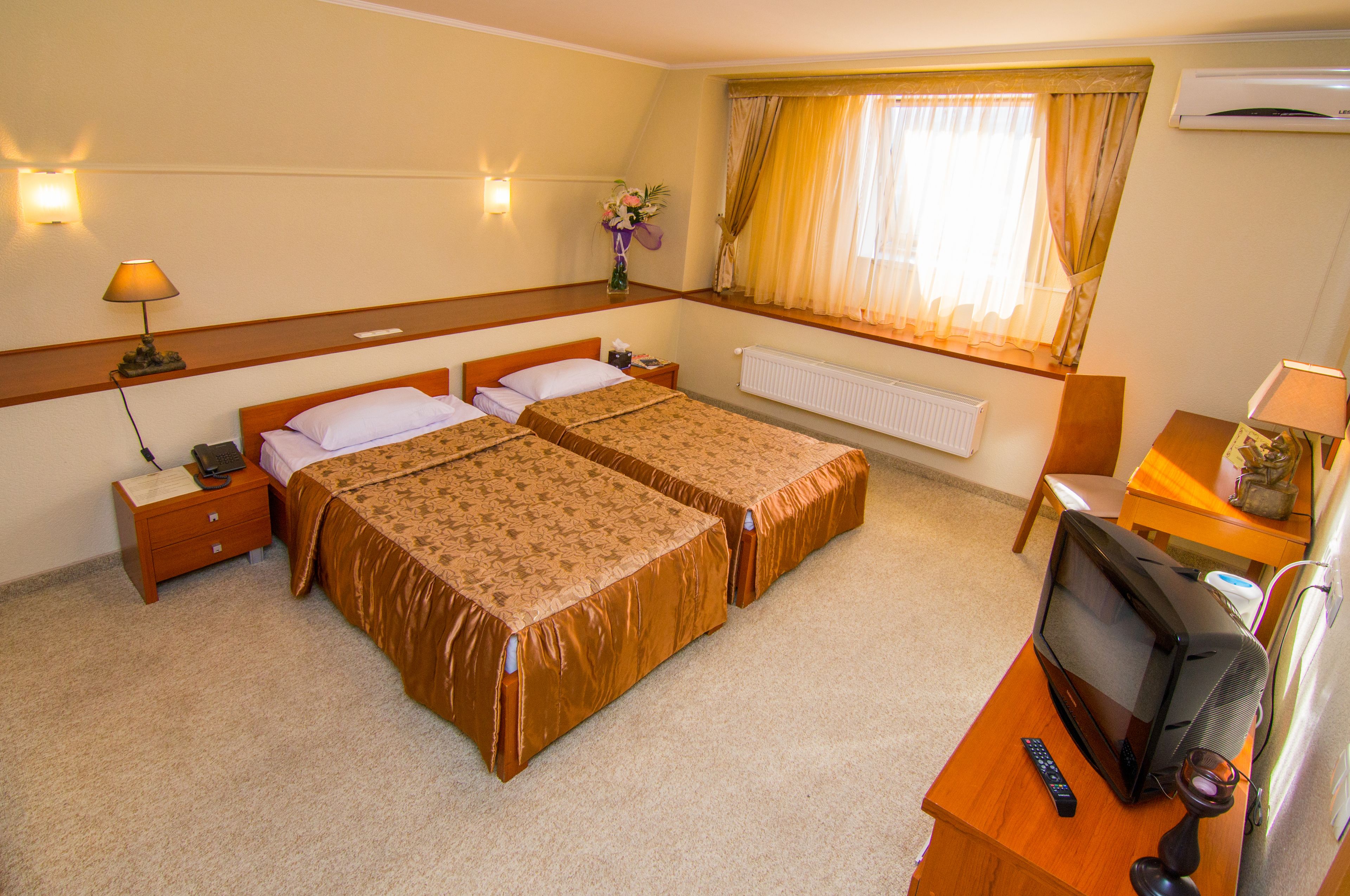 Habitación de hotel con dos camas, un escritorio con televisor, una mesita de noche, una lámpara y una ventana con cortinas.