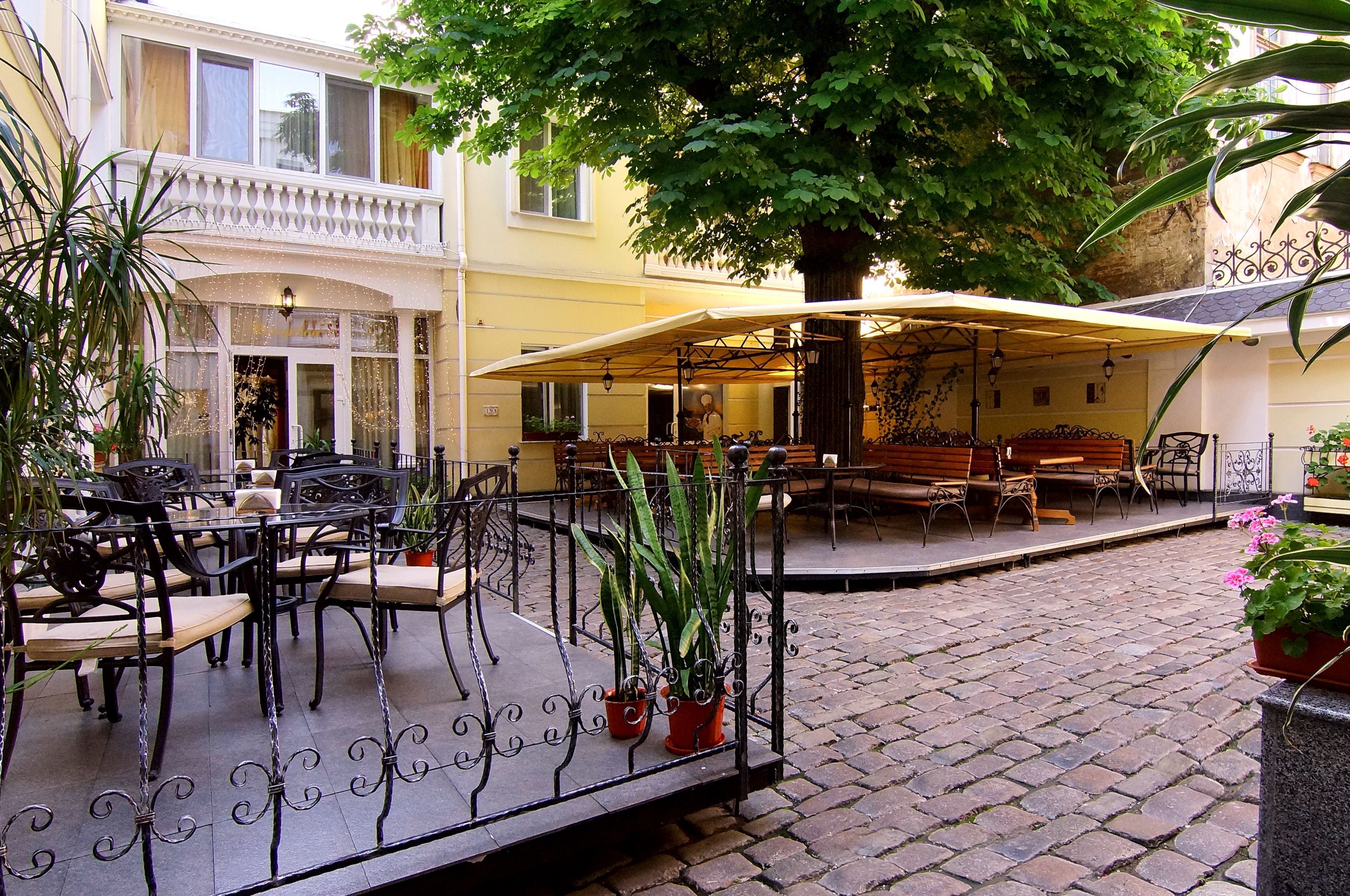 An outdoor café area with tables and chairs, surrounded by potted plants and a large tree providing shade.