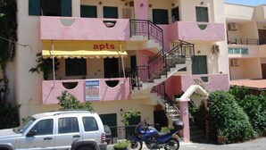 A multi-story building with a pink facade, labeled "Maria Anna Apts" for rent. It features a yellow awning, a staircase, and a parked SUV. - Marianna Hotel Apartments (Malevizi)