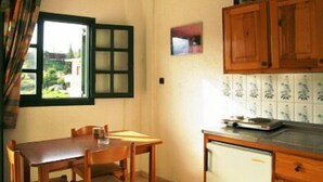 A small kitchen with a table and chairs, a window with green shutters, and wooden cabinets. - Marianna Hotel Apartments (Malevizi)