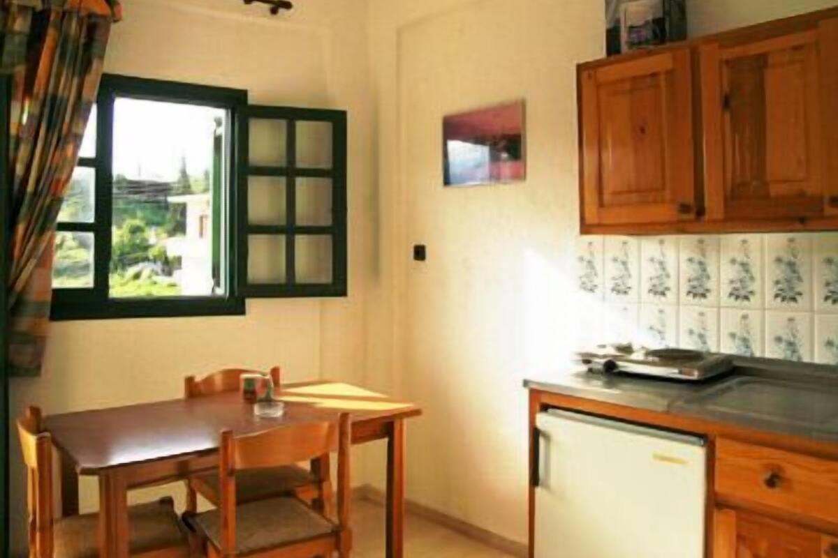 Een kleine keuken met een tafel en stoelen, een raam met groene luiken en houten keukenkastjes.