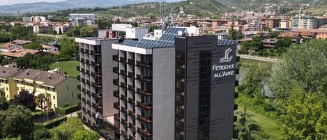 Un edificio moderno con un letrero que dice "RESIDENCE ALL'ADIGE". Cuenta con varios pisos con balcones, rodeado de vegetación y un paisaje montañoso.