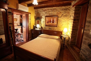 Un dormitorio tradicional con cama de madera, paredes de piedra, un armario de madera y una pequeña mesa con lámpara.