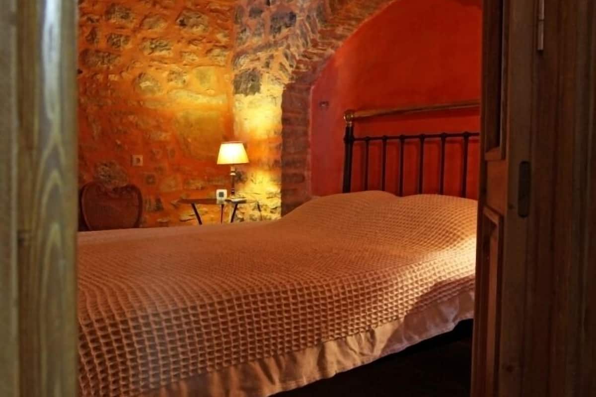 A bedroom with a wooden ceiling, stone walls, a bed with a patterned bedspread, a nightstand, and a lamp.