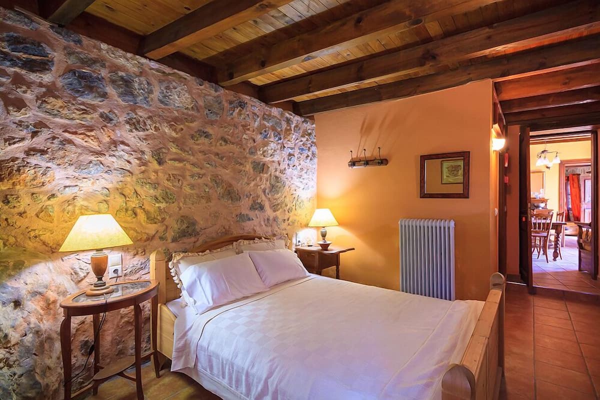 A bedroom with a stone wall, wooden beams, a bed with white linens, two bedside lamps, and a radiator.