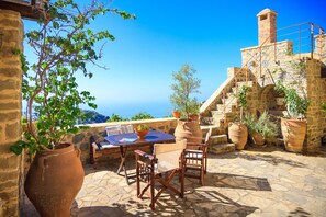 A stone patio with a table and chairs, potted plants, and a view of the sea. - Dorovinis Country Houses (Ierapetra)