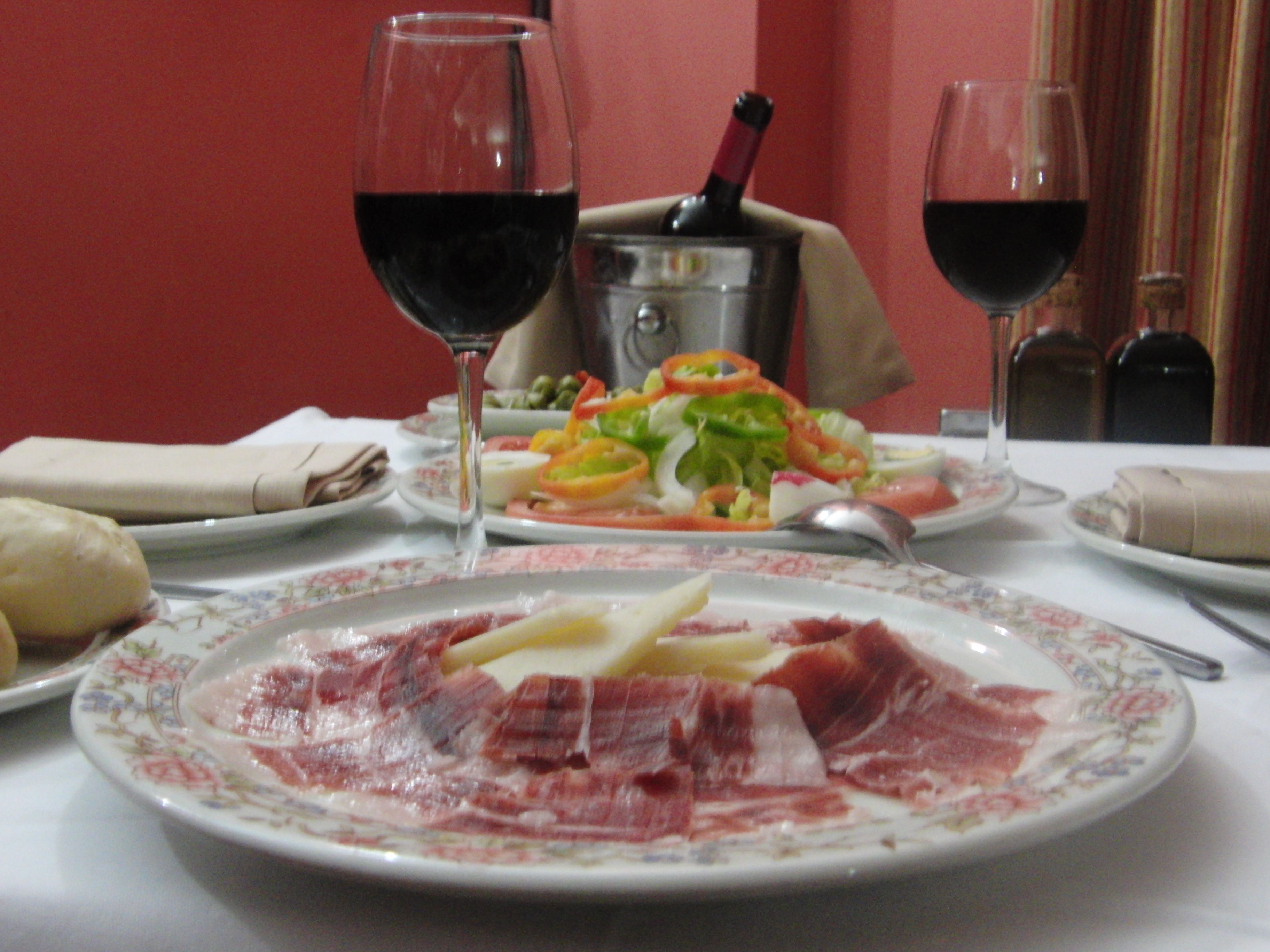 Una mesa de comedor con platos de comida, dos copas de vino tinto y una botella de vino.