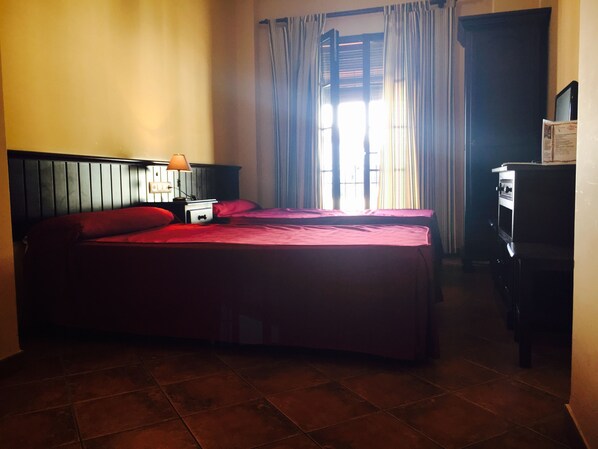 A hotel room with a bed, a nightstand, a lamp, a window with curtains, and a refrigerator.