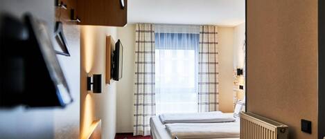 Habitación de hotel con cama, escritorio con computadora, ventana con cortinas y radiador.