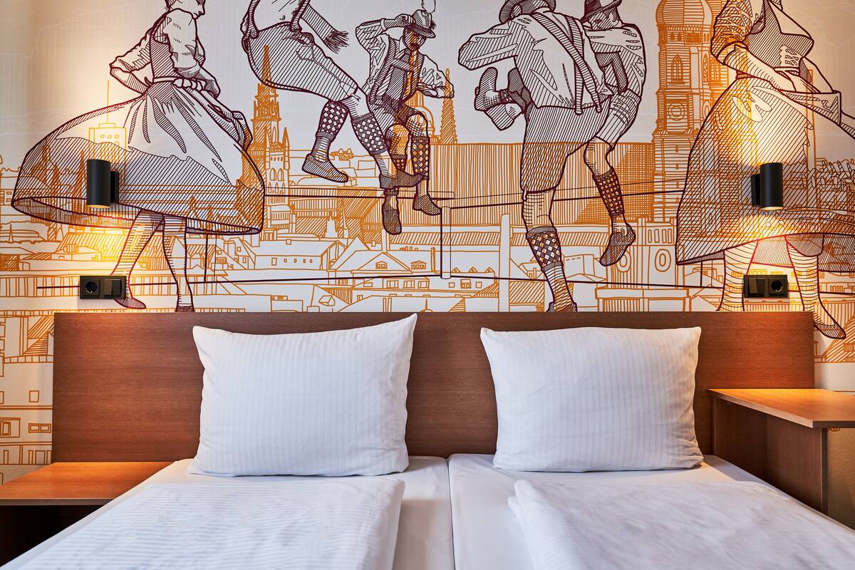 A hotel room with a bed, two pillows, a wooden headboard, and a wall mural depicting historical figures and a cityscape.