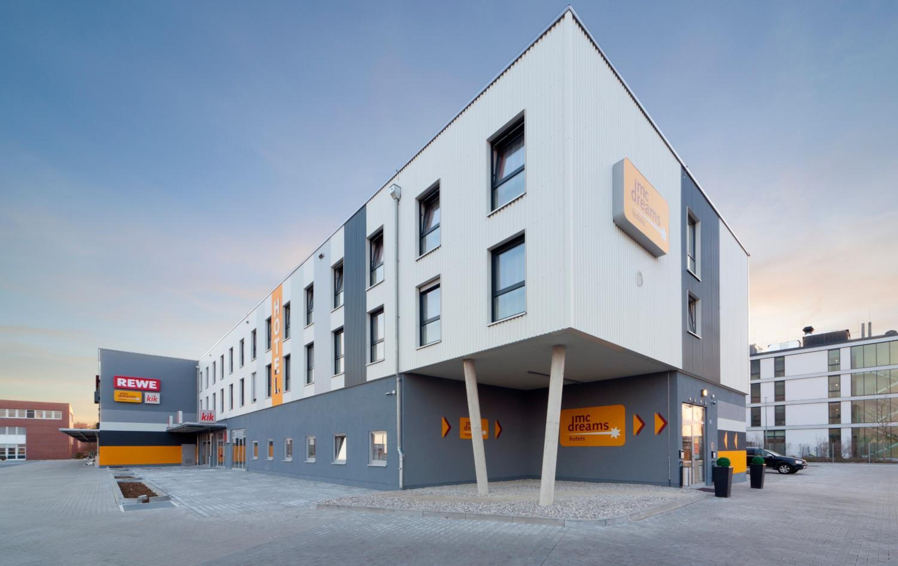 Un edificio moderno di un hotel con tetto piatto, numerose finestre e un cartello con i nomi 'REWE' e 'ikik' sulla sinistra.