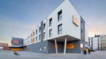 Un edificio moderno di un hotel con tetto piatto, numerose finestre e un cartello con i nomi 'REWE' e 'ikik' sulla sinistra.