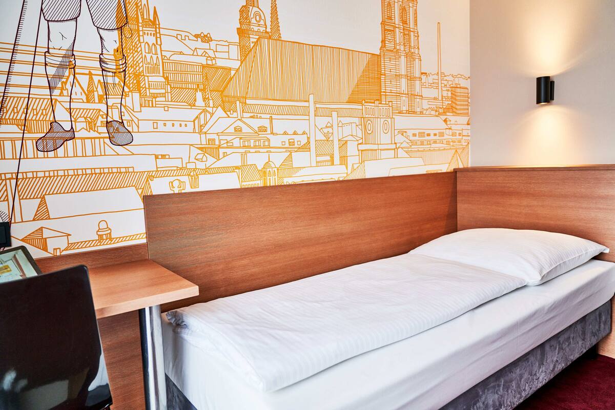 A hotel room with a wooden headboard, a single bed with white bedding, a desk with a chair, and a wall mural of a cityscape.