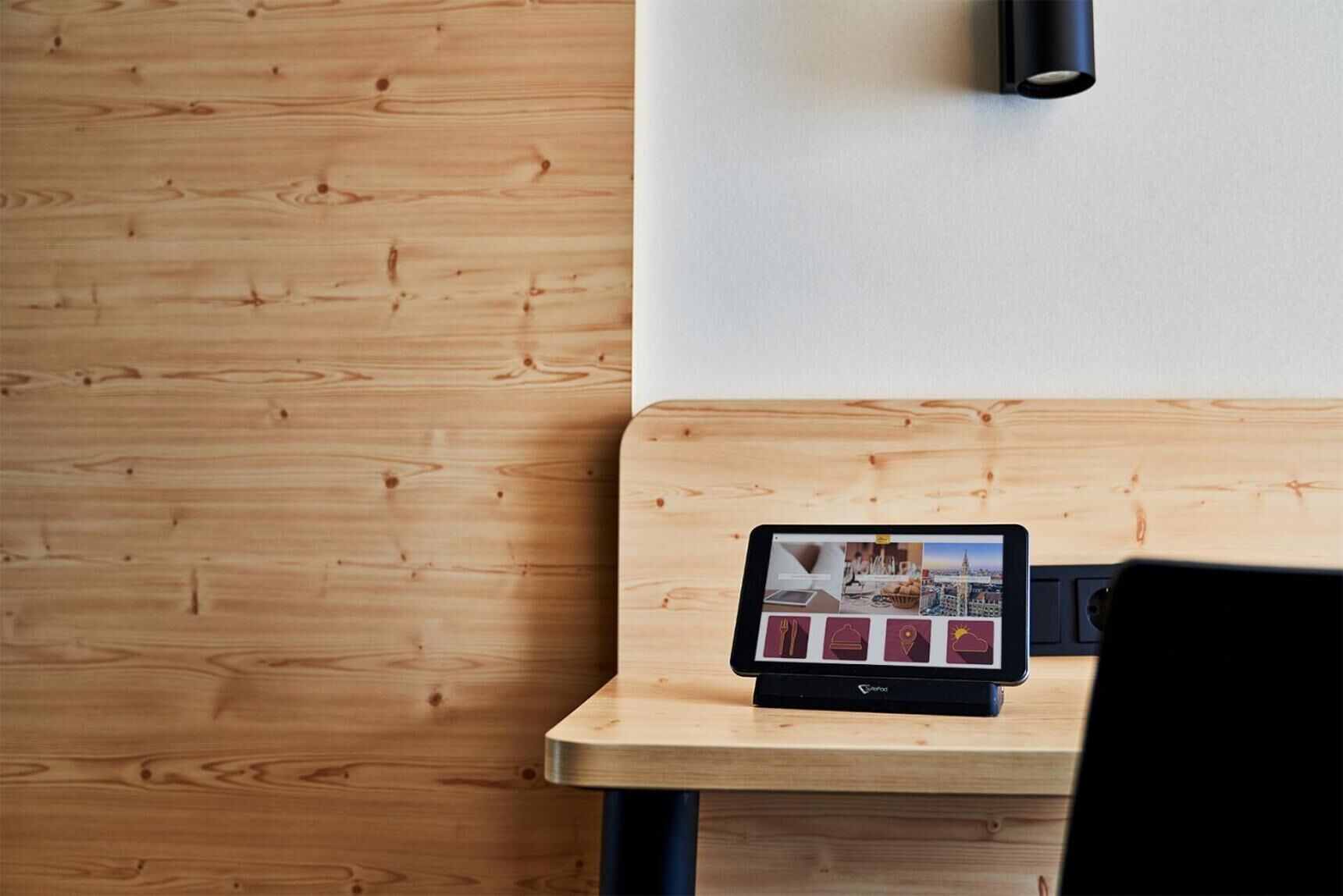 A tablet on a wooden table displaying a cityscape and icons, with a black wall-mounted lamp above.